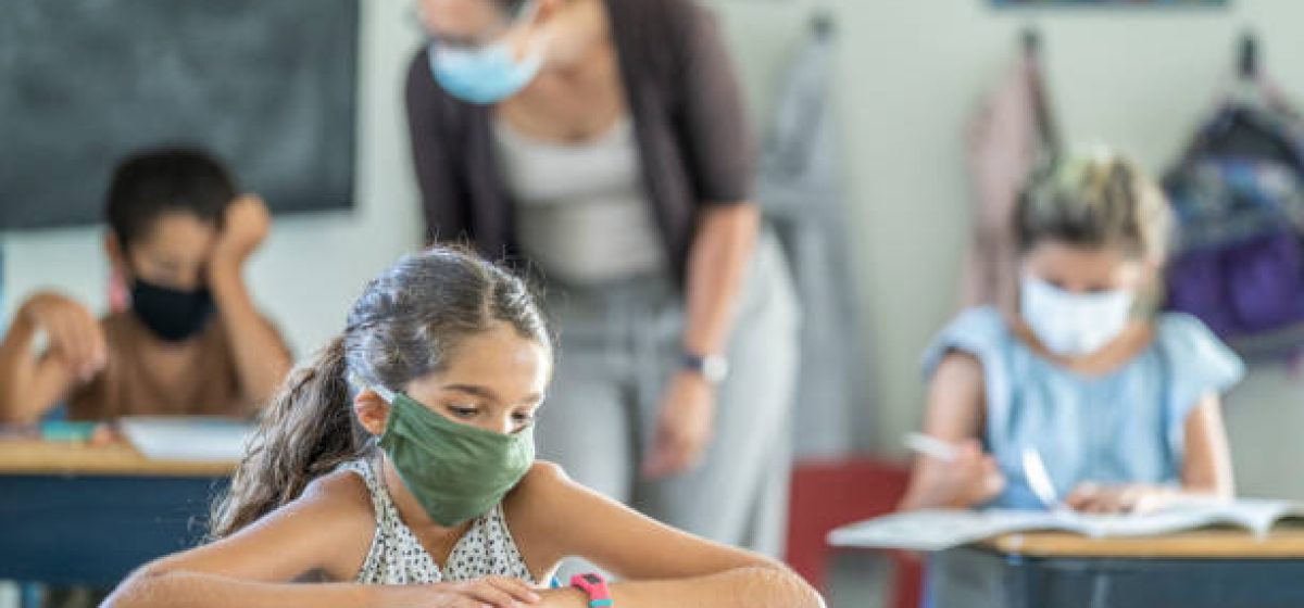12 year old girl wearing a reusable, protective face mask in classroom while working on school work at her desk.