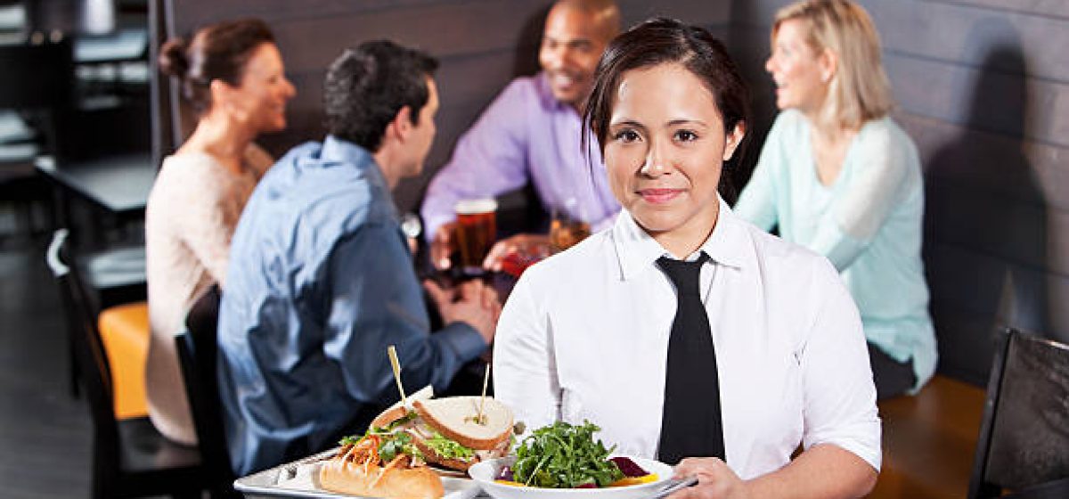 Hispanic waitress (20s) carrying tray with food.  Multi-ethnic customers sitting in background talking and drinking.