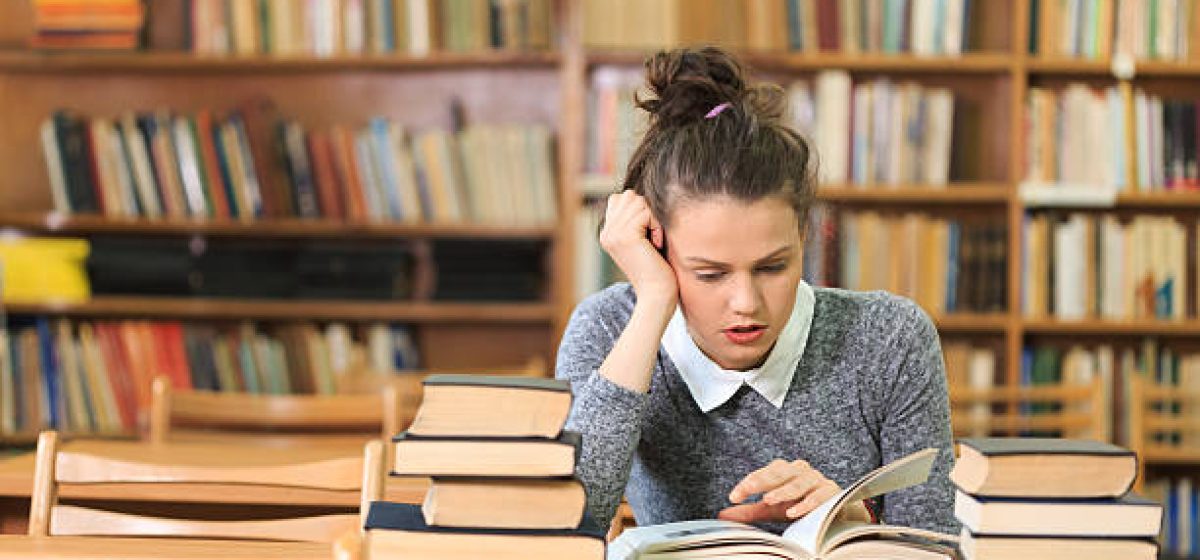 Concentrated young woman sitting on desk and reading a book in the library. Hand on cheek and gray pullover reading a book on desk. Bokshelves as background, stack of books in foreground.